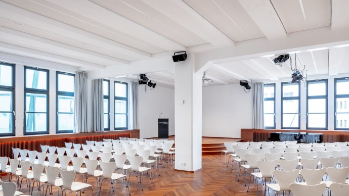 Modern hall with white chairs, wooden floor and stage. Large windows let in plenty of light. Lighting and loudspeakers on the ceiling., &copy; &copy; markus guhl fotografie