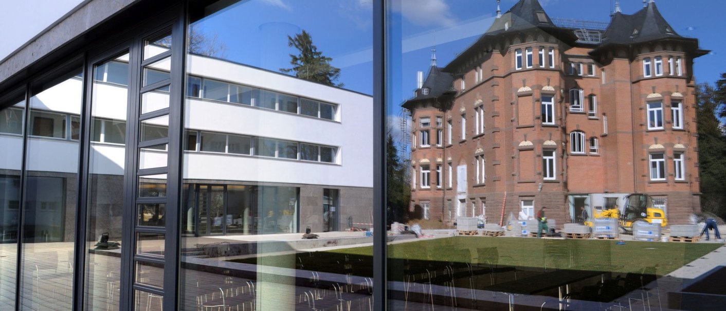 Modern glass fa&ccedil;ade of the Evangelical Academy Bad Boll reflects a historic building and the blue sky. Chairs can be seen in the foreground., &copy; Eabb_Giacinto_Carlucci