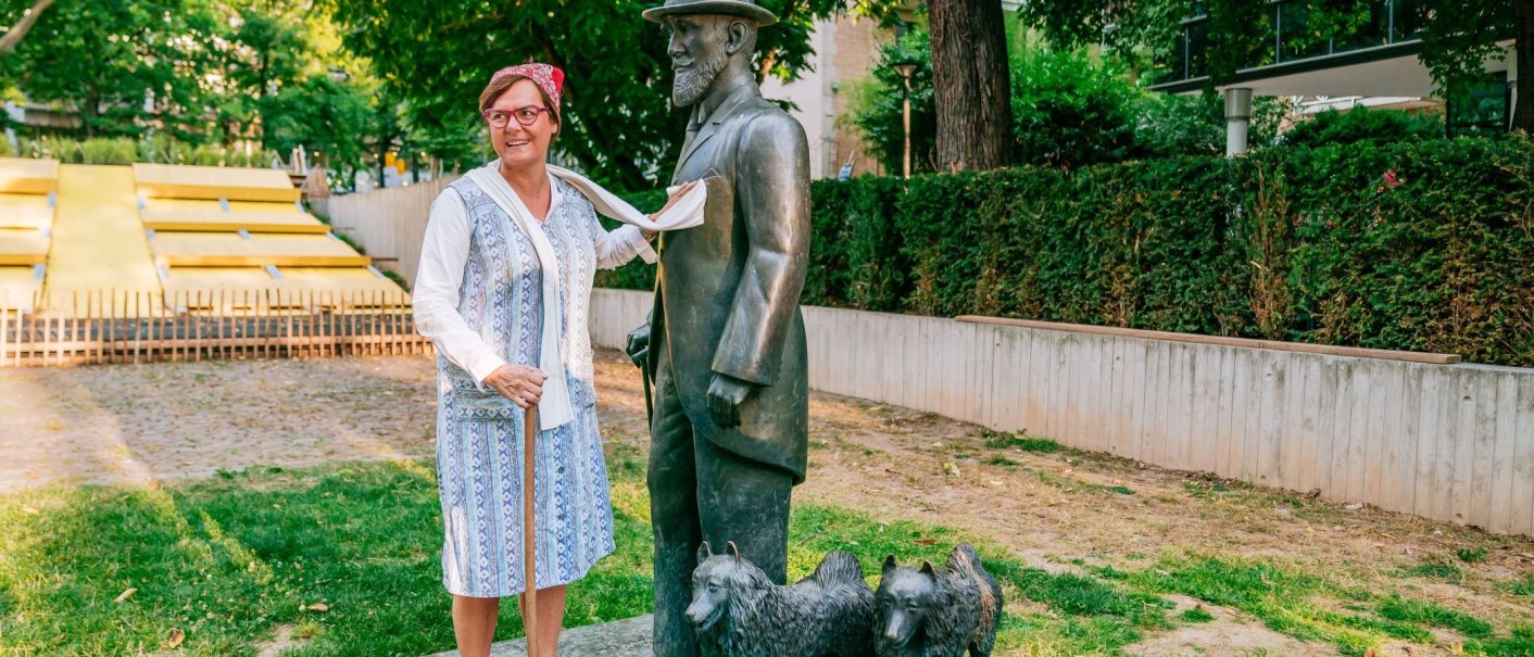 Frau in traditioneller Kleidung posiert lächelnd neben einer Bronzestatue eines Mannes mit Hut und zwei Hunden im Freien., © Thomas Niedermüller Frau in traditioneller Kleidung posiert lächelnd neben einer Bronzestatue eines Mannes mit Hut und zwei Hunden im Freien., © Thomas Niedermüller