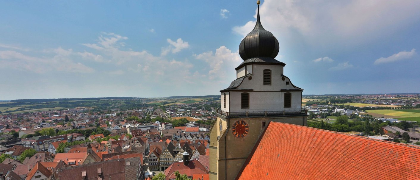 View of Herrenberg with a striking church tower in the foreground and a wide cityscape in the background under a blue sky., © Stadt Herrenberg View of Herrenberg with a striking church tower in the foreground and a wide cityscape in the background under a blue sky., © Stadt Herrenberg