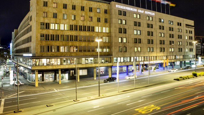 Nachtansicht des Steigenberger Graf Zeppelin Hotels in Stuttgart. Die beleuchtete Fassade zeigt mehrere Fenster und wehende Flaggen auf dem Dach., © Steigenberger Hotels AG, Tanja Ganzer