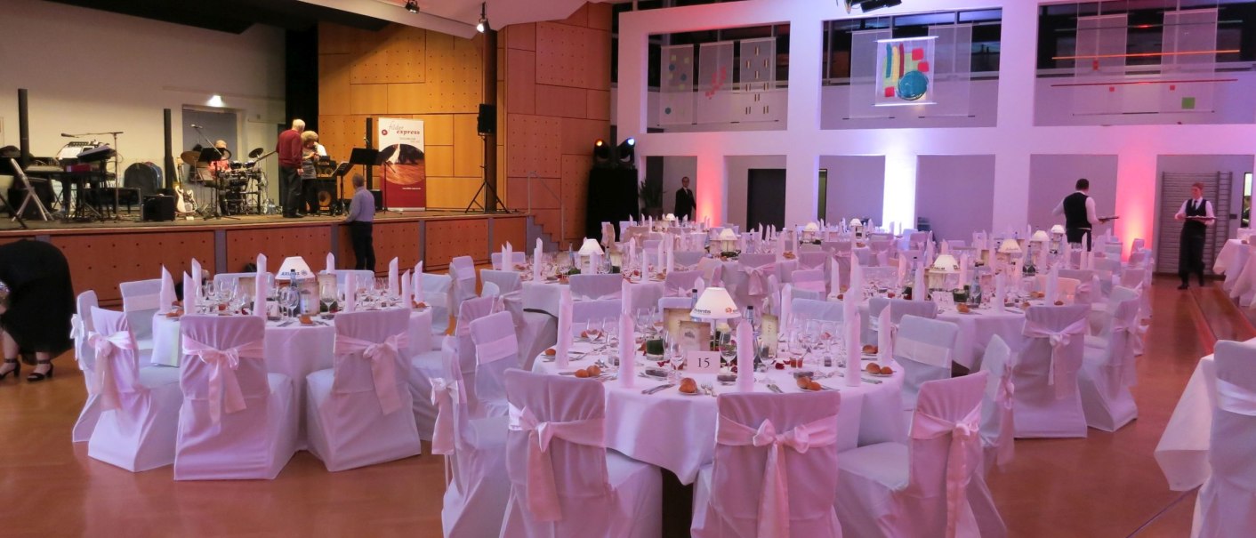 Ein festlich dekorierter Saal mit runden Tischen, weißen Stuhlhussen und einer Bühne. Die Beleuchtung ist warm und einladend., © GemeindeBaltmannsweiler_GF Ein festlich dekorierter Saal mit runden Tischen, weißen Stuhlhussen und einer Bühne. Die Beleuchtung ist warm und einladend., © GemeindeBaltmannsweiler_GF