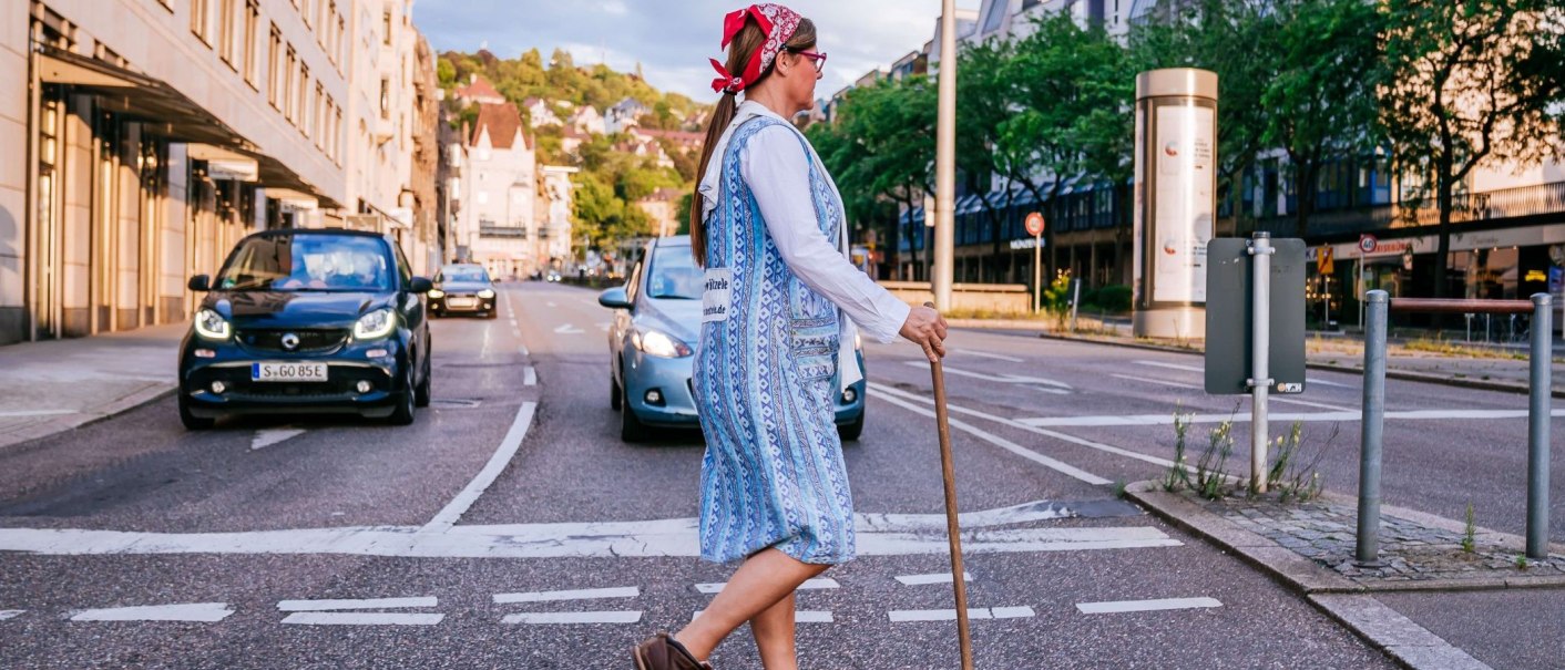 Eine Frau mit Kopftuch und Besen überquert eine Straße in einer Stadt. Autos sind im Hintergrund zu sehen., © Thomas Niedermüller Eine Frau mit Kopftuch und Besen überquert eine Straße in einer Stadt. Autos sind im Hintergrund zu sehen., © Thomas Niedermüller