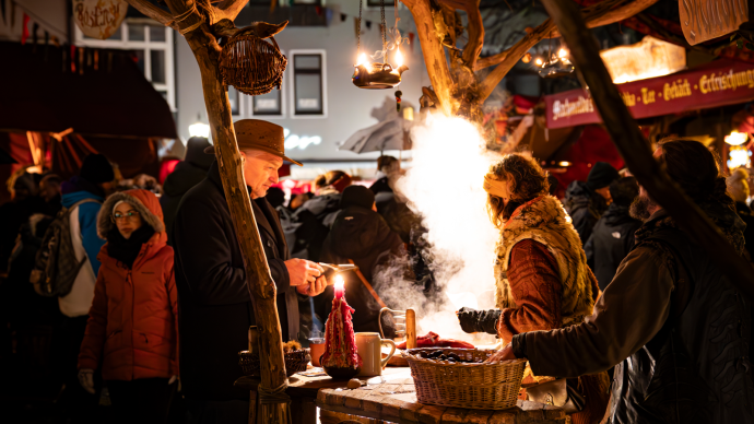 Lively Christmas market with people standing at a stall with candles and steaming food. Festive lighting and a bustling atmosphere., © Stuttgart-Marketing GmbH, Sarah Schmid