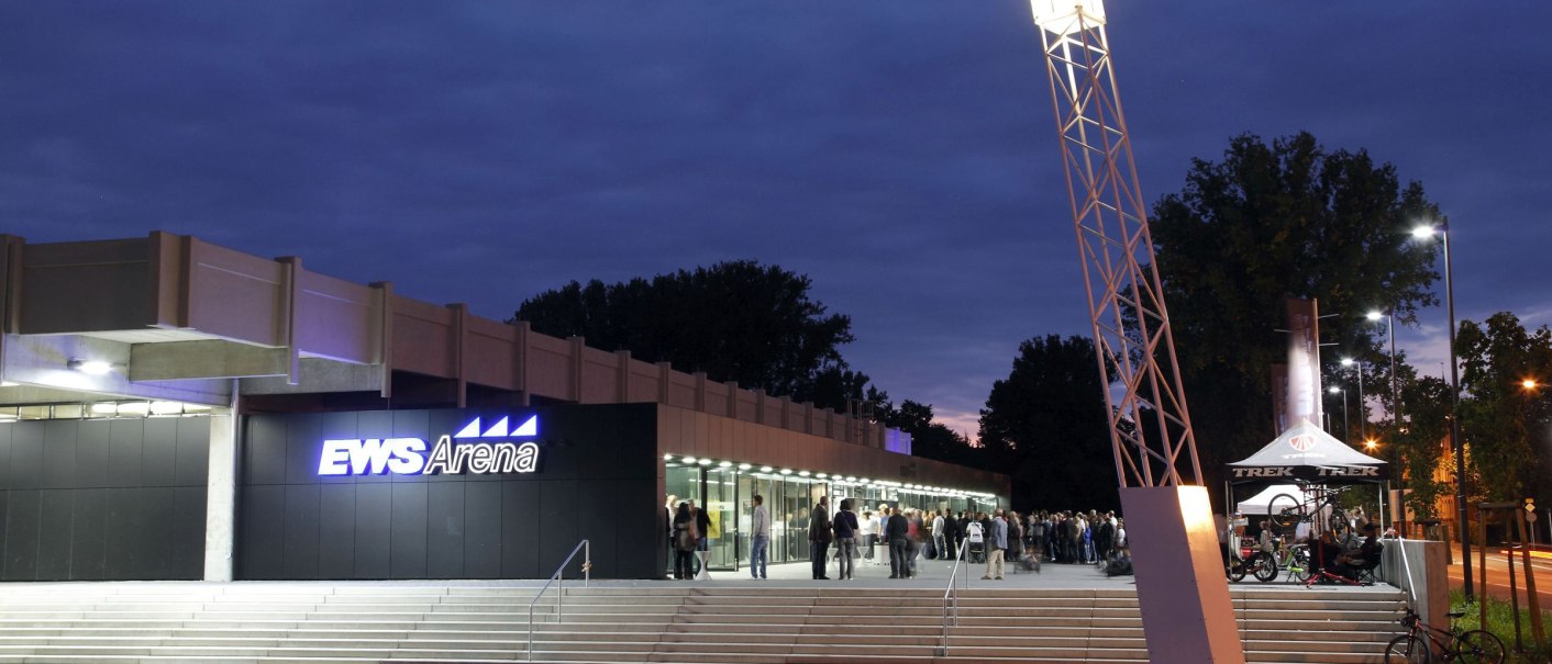 The EWS Arena at dusk, people standing in front of the entrance. An illuminated pole and a bicycle stand are visible., © Dietmar Strauss The EWS Arena at dusk, people standing in front of the entrance. An illuminated pole and a bicycle stand are visible., © Dietmar Strauss