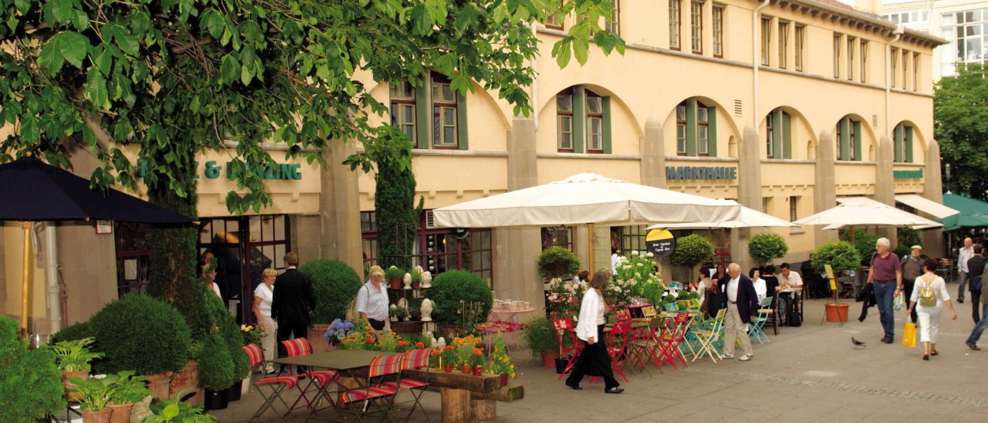 Au&szlig;enbereich der Stuttgarter Markthalle mit Tischen, St&uuml;hlen und Pflanzen. Menschen flanieren und sitzen unter Sonnenschirmen. Geb&auml;ude im Hintergrund., &copy; Stuttgart-Marketing GmbH