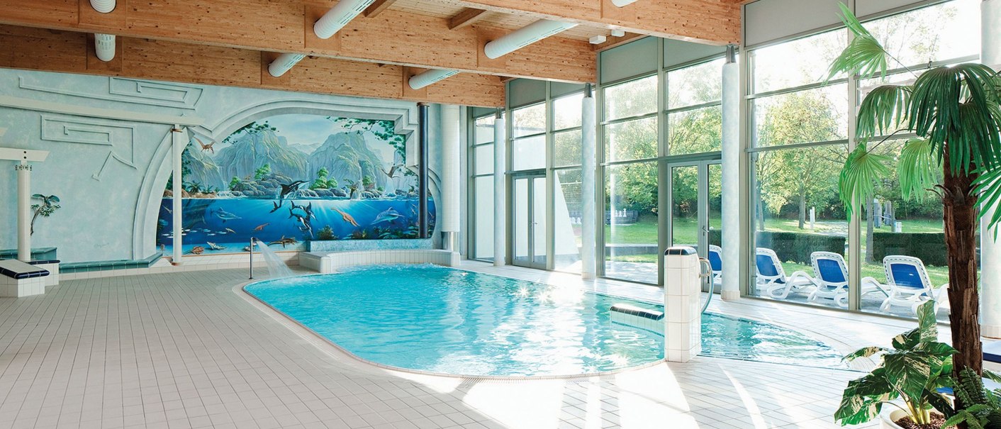 Heller Innenpoolbereich im Seminaris Hotel Bad Boll mit Wandgemälde, Liegestühlen und Pflanzen. Große Fenster lassen viel Licht herein., © Seminaris Hotels