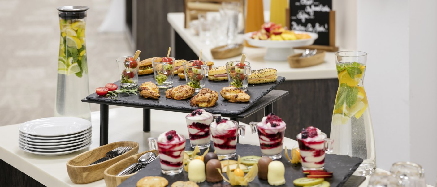 Ein Buffet mit verschiedenen Snacks wie Geb&auml;ck, Desserts und Obst. Im Hintergrund stehen Karaffen mit Wasser und Saft sowie eine Tafel mit Men&uuml;beschreibung., &copy; ARCOTEL Hotels