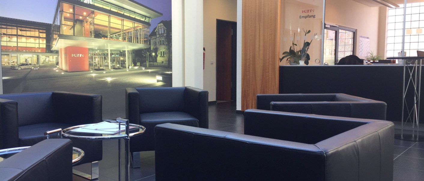 Modern foyer with black leather armchairs, a round table and a large mural of the K3N building. Reception area in the background., © k3nmaisch