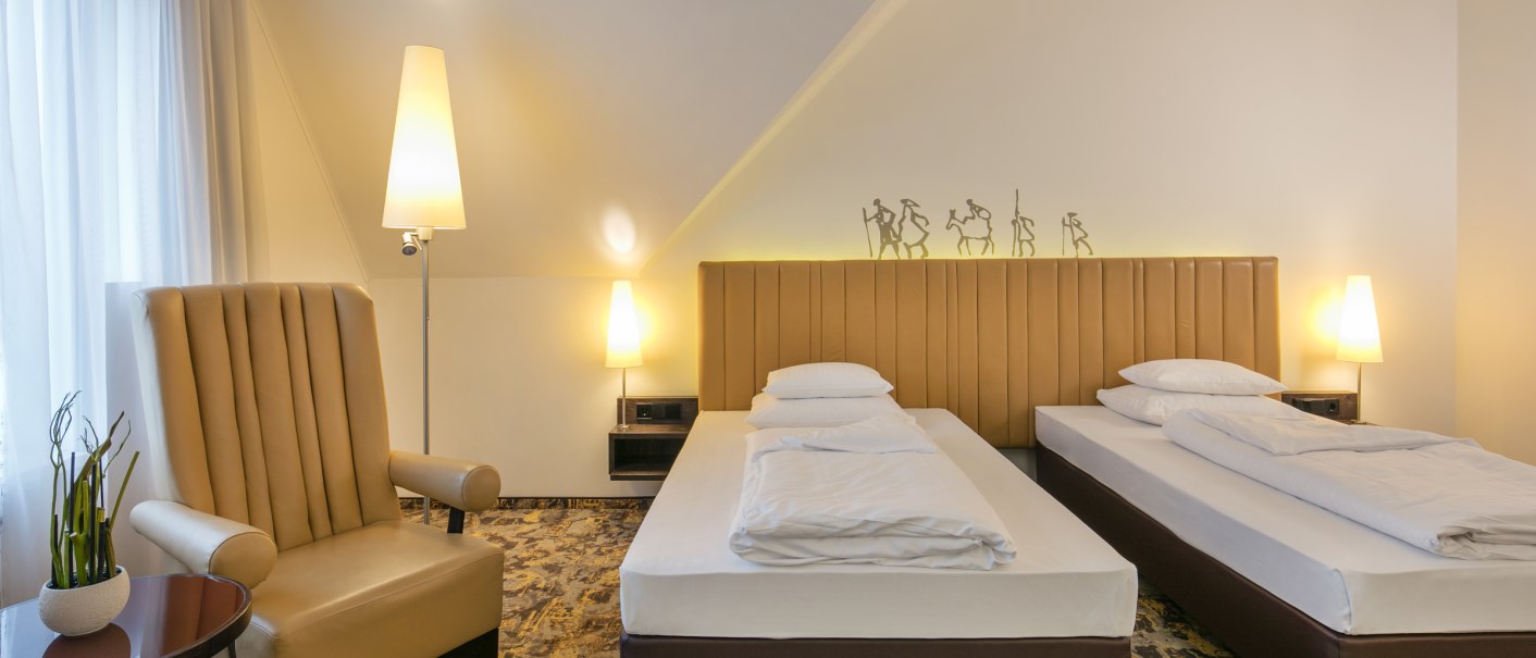 Hotelzimmer mit zwei Einzelbetten, einem beigen Sessel, Stehlampen und dekorativen Figuren an der Wand. Helle, gemütliche Atmosphäre., © ARCOTEL Hotels