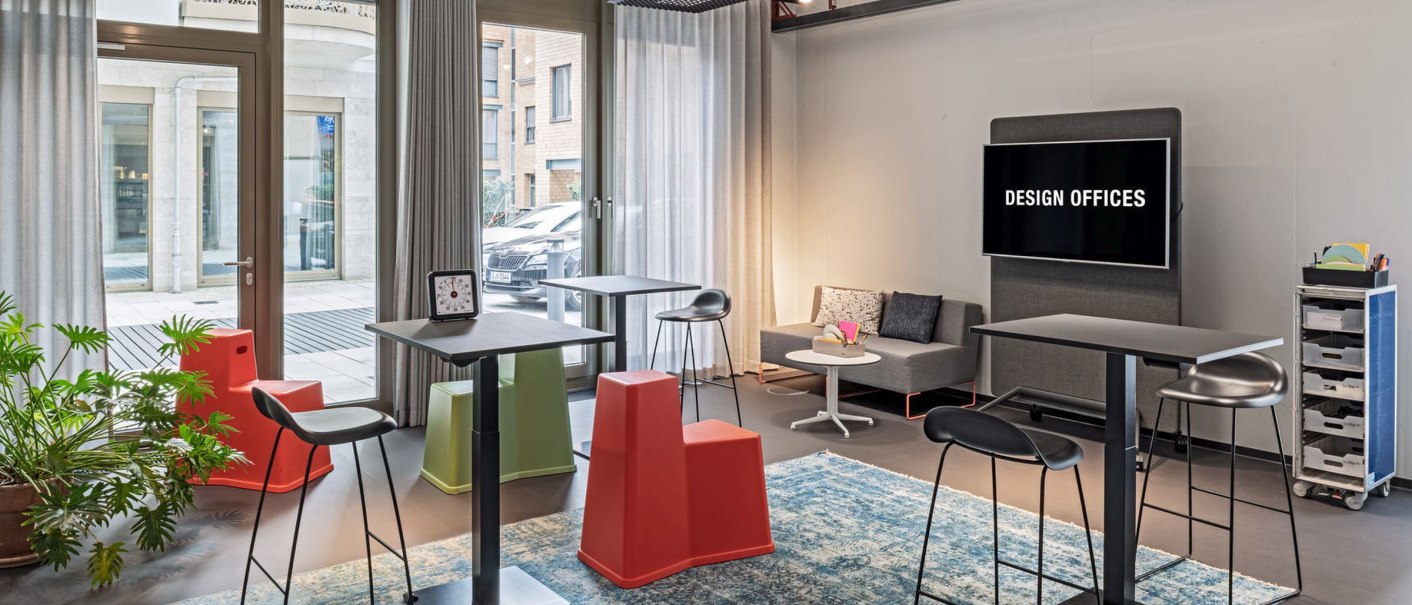 Moderner Raum mit Stehtischen, bunten Hockern, Sofa und Bildschirm. Große Fenster, Pflanzen und ein blauer Teppich schaffen eine einladende Atmosphäre., © Design Offices