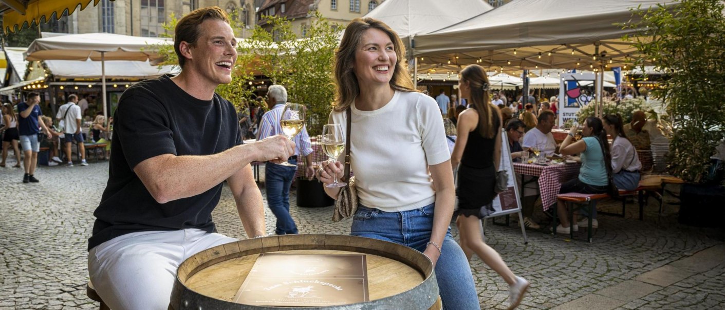 Zwei Personen sto&szlig;en mit Weingl&auml;sern auf dem Stuttgarter Weindorf an. Im Hintergrund sind St&auml;nde und Besucher zu sehen., &copy; Stuttgart-Marketing GmbH, Sarah Schmid