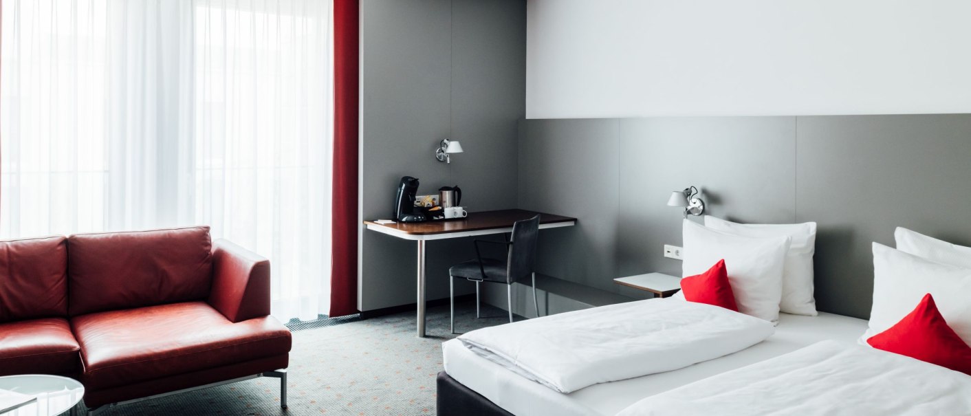 Modernes Hotelzimmer mit Doppelbett, rotem Sofa, Schreibtisch und Kaffeemaschine. Helle Vorhänge und rote Akzente schaffen eine einladende Atmosphäre., © VHE Mo.Stuttgart