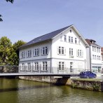 White building on a river with a bridge, surrounded by trees and other buildings under a blue sky., &copy; econvent