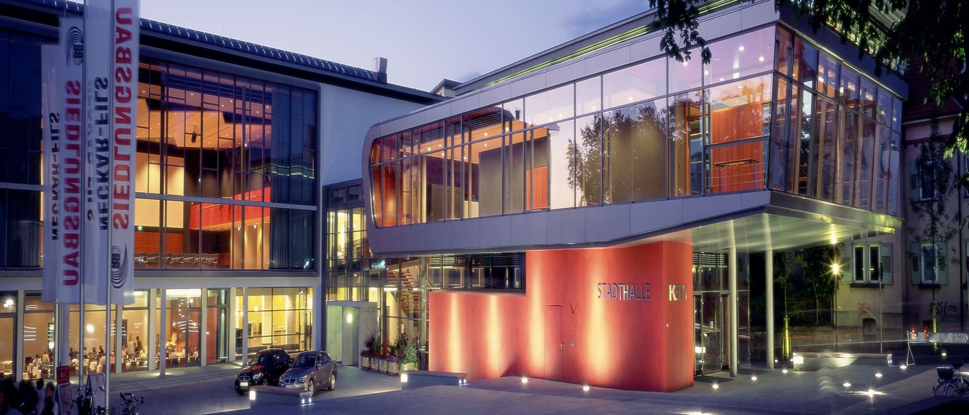 The Stadthalle K3N at dusk, modern glass architecture with illuminated forecourt and flagpoles., © k3nmaisch