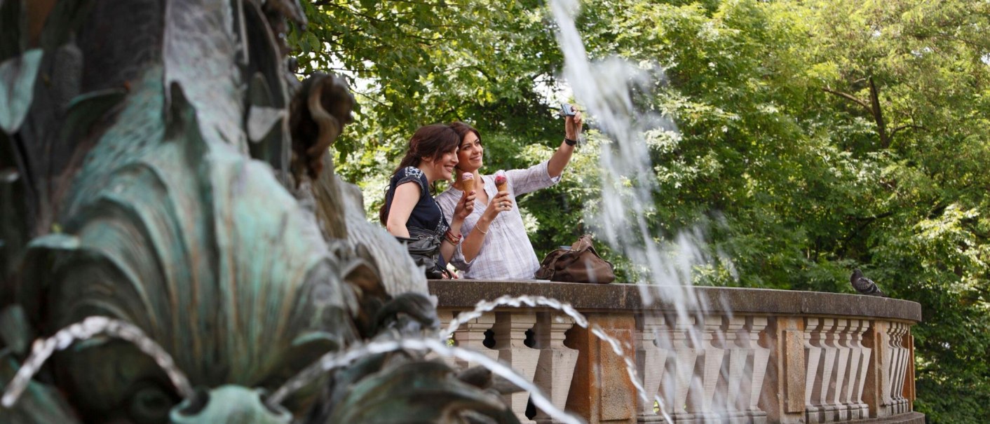 Zwei Frauen machen ein Selfie am Galateabrunnen, halten Eis in der Hand. Im Hintergrund sind Bäume und ein Wasserspritzer zu sehen., © Stuttgart-Marketing GmbH Christoph Düpper