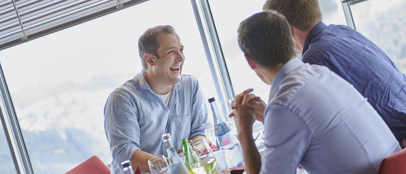 Three men sit laughing at a table in a restaurant with large windows. There are bottles of water and glasses on the table., © Martin Sigmund Three men sit laughing at a table in a restaurant with large windows. There are bottles of water and glasses on the table., © Martin Sigmund