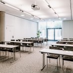 Moderner Tagungsraum mit Tischen und St&uuml;hlen, einem Deckenprojektor und gro&szlig;en Fenstern mit Jalousien. Helle, neutrale Farben dominieren den Raum., &copy; VHE Mo.Stuttgart