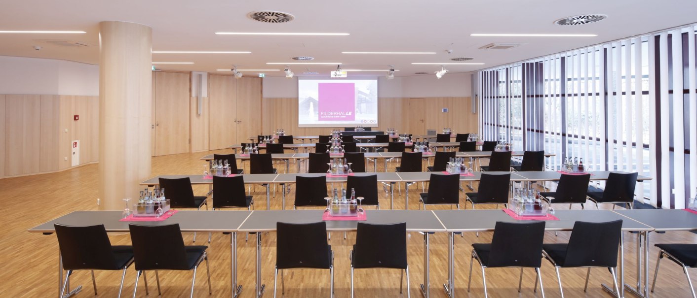 Modern conference room with rows of tables and chairs, equipped with water bottles and glasses. Light wooden floors and large windows., © Kristof Lange