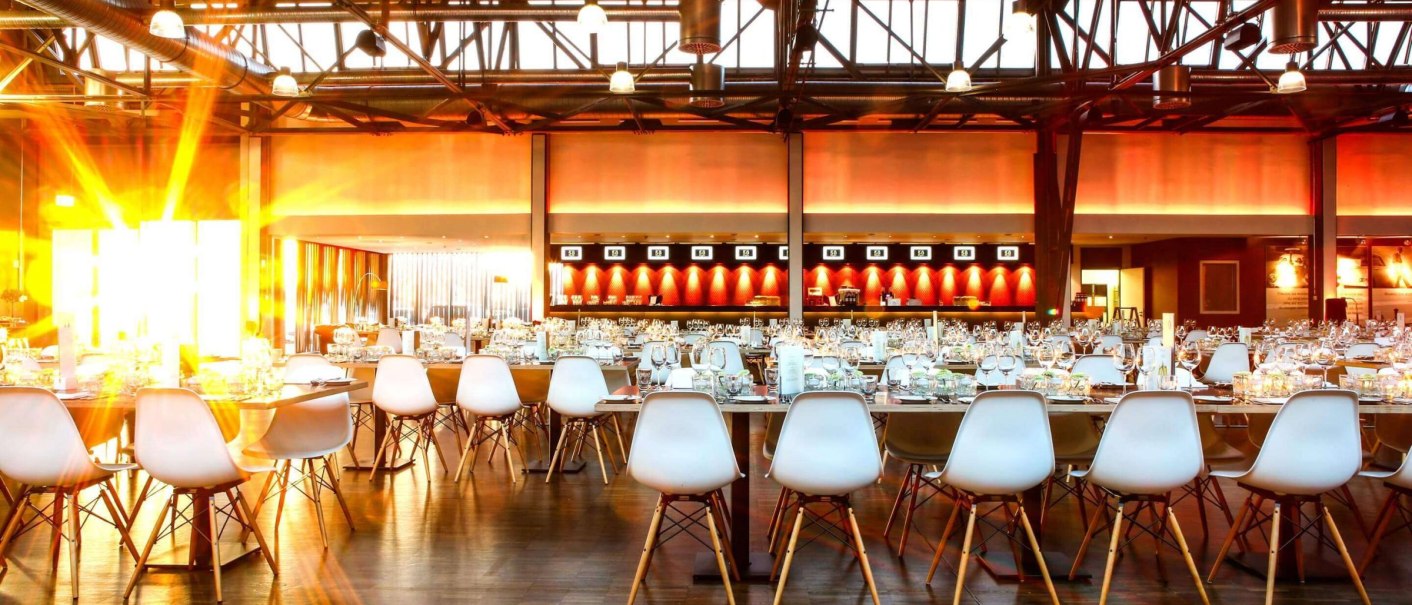 Ein großer Saal mit festlich gedeckten Tischen und weißen Stühlen. Sonnenlicht strömt durch die Fenster und beleuchtet den Raum warm., © rauschenbergercatering