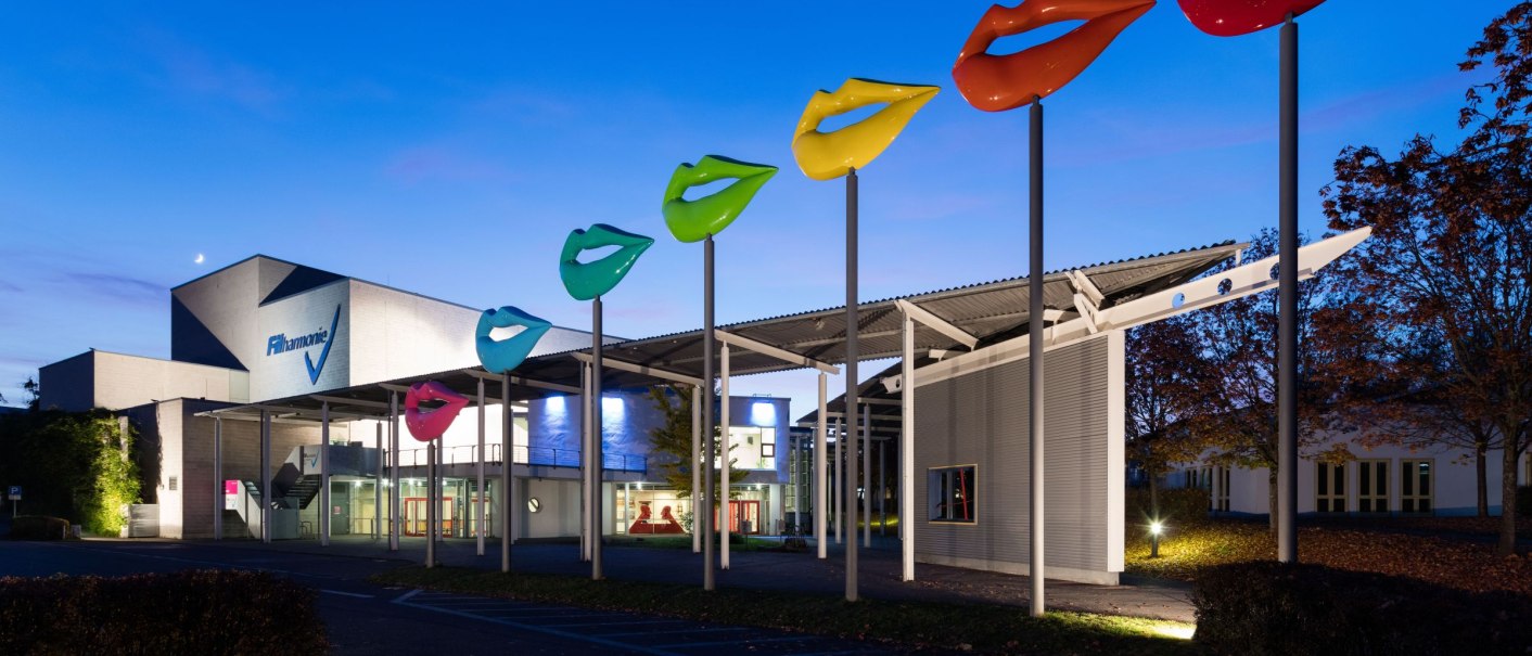 Die FILharmonie bei Dämmerung, mit bunten Lippen-Skulpturen auf hohen Stangen vor dem Gebäude., © Filharmonie_gf Die FILharmonie bei Dämmerung, mit bunten Lippen-Skulpturen auf hohen Stangen vor dem Gebäude., © Filharmonie_gf