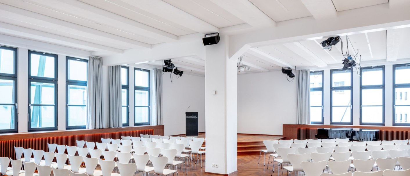 Modern hall with white chairs, wooden floor and stage. Large windows let in plenty of light. Lighting and loudspeakers on the ceiling., © © markus guhl fotografie