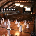 Innenansicht der Stadthalle Herrenberg: Leere Stuhlreihen, Stehtische mit weißen Tischdecken, Bühne mit Projektor, Holzboden und hohe Decke., © Stadt Herrenberg