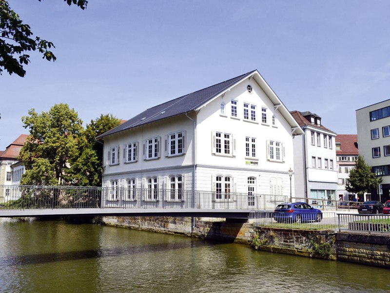 Weißes Gebäude an einem Fluss mit Brücke, umgeben von Bäumen und weiteren Gebäuden unter blauem Himmel., © econvent
