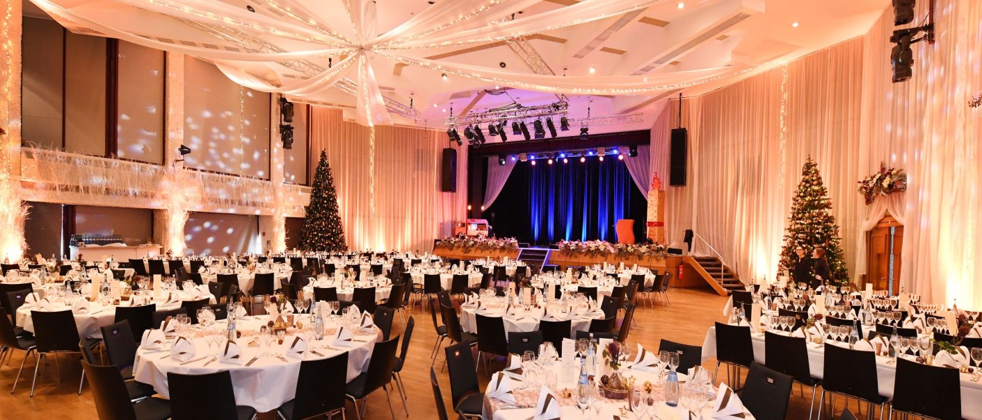 Large hall in the Filderhalle, festively decorated with round tables, white tablecloths and Christmas trees. Stage with blue lighting in the background., © Guenter E. Bergmann - Photography