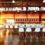 A large hall with festively laid tables and white chairs. Sunlight streams through the windows and illuminates the room warmly., © rauschenbergercatering