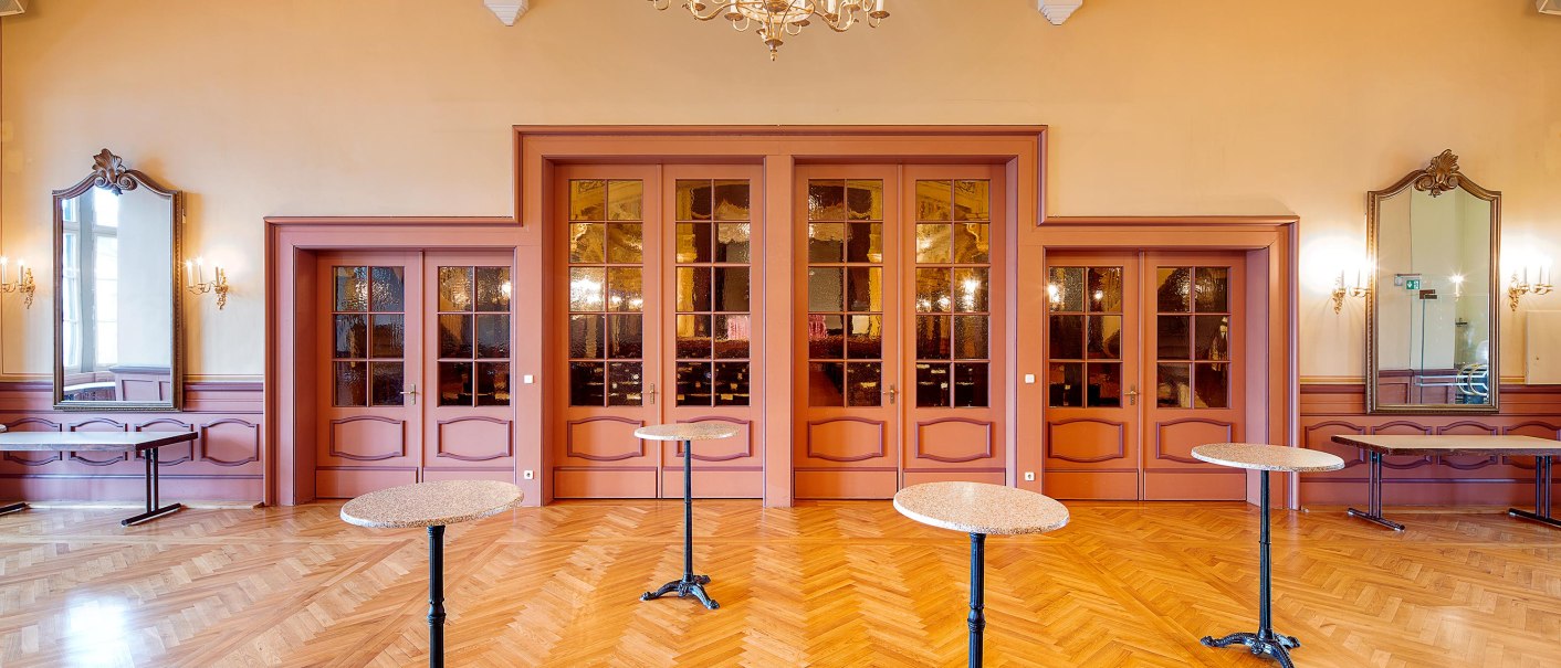 Elegant hall with parquet floor, chandelier and round bar tables. Large mirrors and ornate wall lights adorn the walls., © Stauch