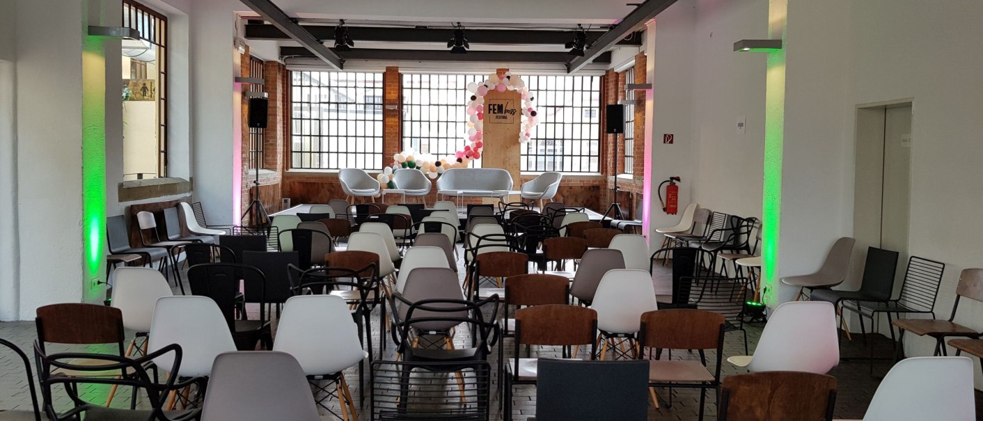 An empty event room with rows of chairs and a stage, decorated with colorful balloons and the lettering 'FEM'. Large windows in the background., &copy; Julian Rettig