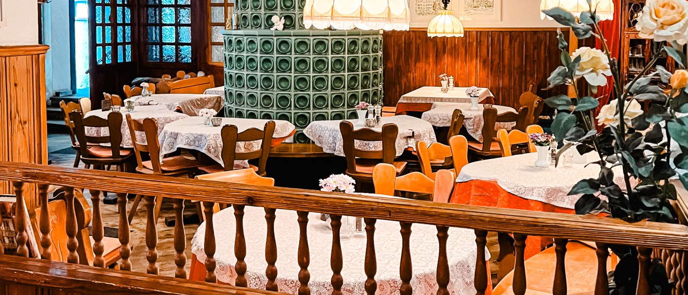 Cozy dining room with wooden furniture, a green tiled stove and decorative tablecloths. Hanging lamps and flower arrangements complete the ambience., © Weinstube Kachelofen
