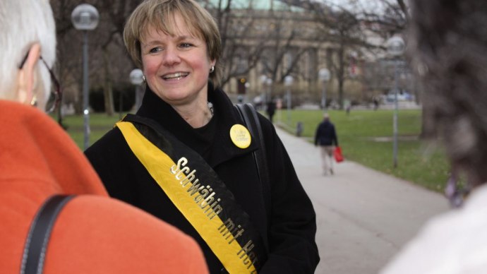 Eine Frau mit einer Schärpe 'Schwäbin mit Herz' lächelt und spricht mit anderen Personen in einem Park., © Stuttgart-Marketing GmbH Eine Frau mit einer Schärpe 'Schwäbin mit Herz' lächelt und spricht mit anderen Personen in einem Park., © Stuttgart-Marketing GmbH