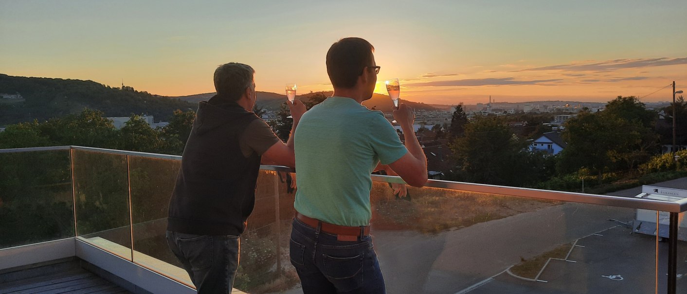 Zwei Personen stehen auf einer Dachterrasse und genießen den Sonnenuntergang mit einem Getränk in der Hand., © Weingaertner-Esslingen-eG Zwei Personen stehen auf einer Dachterrasse und genießen den Sonnenuntergang mit einem Getränk in der Hand., © Weingaertner-Esslingen-eG