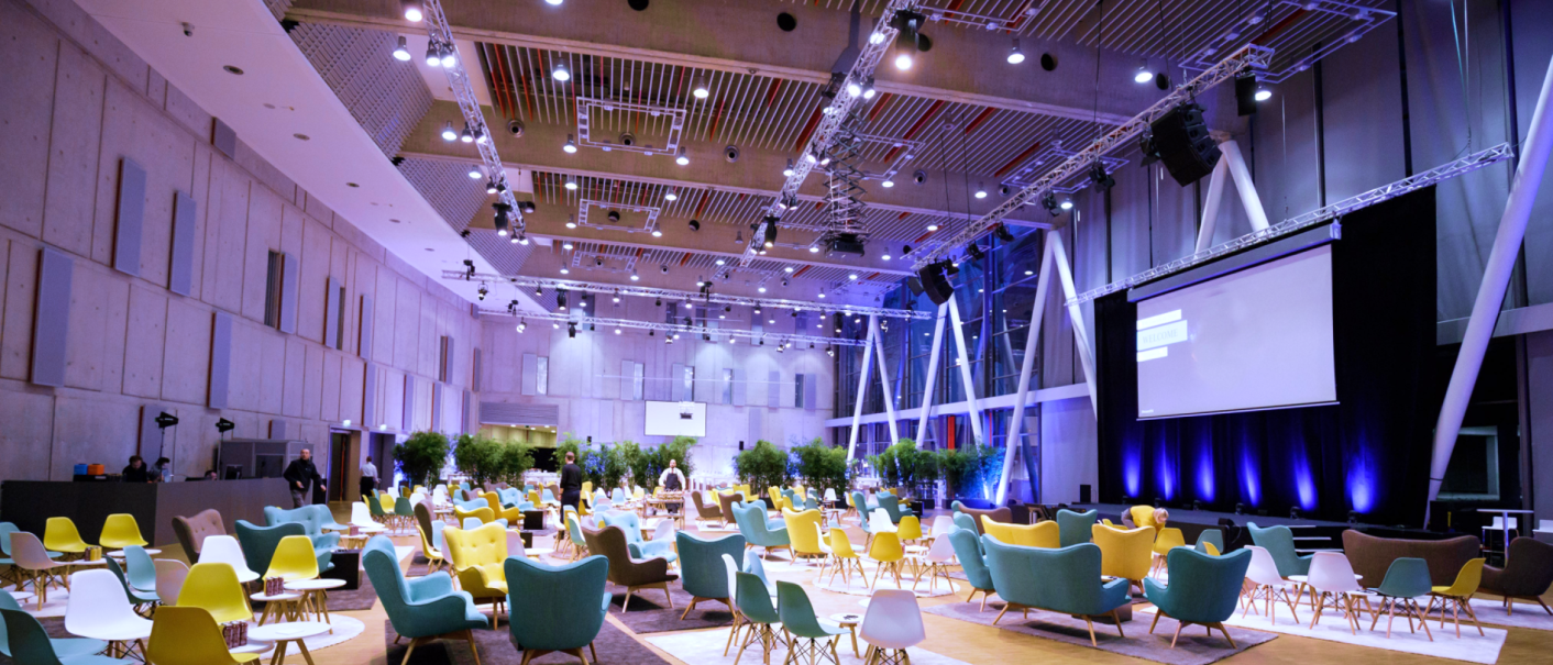 Großer, moderner Raum mit bunten Stühlen und Sofas, Pflanzen und einer großen Leinwand. Die Decke ist mit vielen Lichtern ausgestattet., © Carl Benz Arena