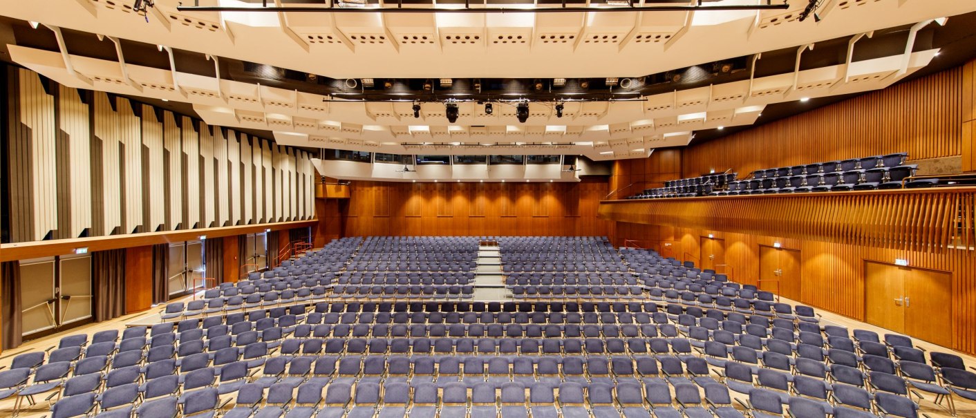 Großer Veranstaltungssaal mit leeren Stuhlreihen und Bühne, moderne Architektur, Holzverkleidung und helle Beleuchtung., © Tobias Fröhner Photography
