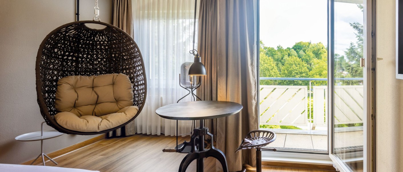 Modernes Hotelzimmer mit Hängesessel, rundem Tisch und Stuhl. Offene Balkontür mit Blick auf grüne Landschaft., © Seminaris Hotels (Gerhard Hinz)