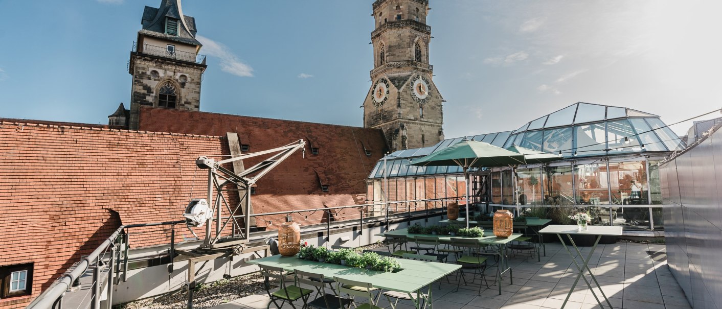 Roof terrace with green tables and chairs, surrounded by glass structures. Two church spires can be seen in the background, under a blue sky., © OutOfOffice GmbH Roof terrace with green tables and chairs, surrounded by glass structures. Two church spires can be seen in the background, under a blue sky., © OutOfOffice GmbH
