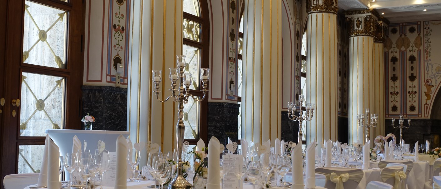 Eleganter Marmorsaal mit hohen, verzierten Säulen, gedeckten Tischen und Kerzenleuchtern. Große Fenster lassen Licht herein., © marmorsaalgmbh