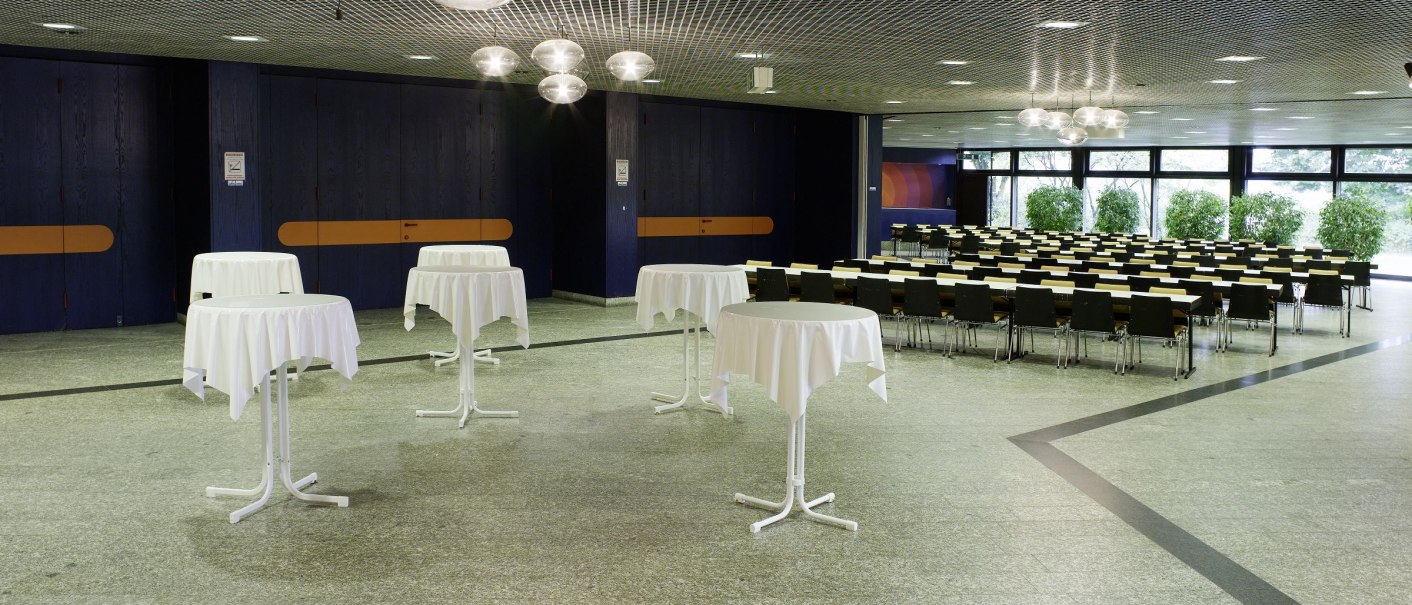 The foyer of the Osterfeldhalle features bar tables with white tablecloths and rows of chairs in front of large windows and plants., © eslive_gf The foyer of the Osterfeldhalle features bar tables with white tablecloths and rows of chairs in front of large windows and plants., © eslive_gf
