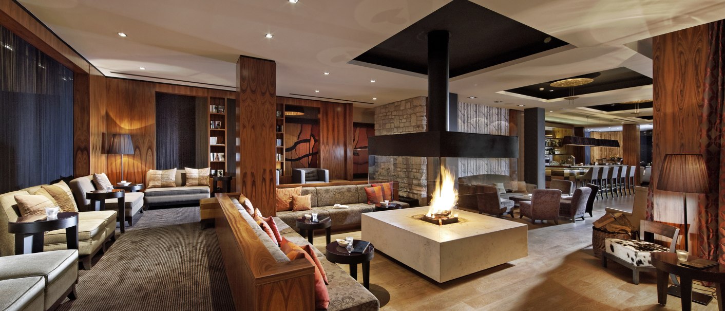 Elegante Lounge mit zentralem Kamin, gemütlichen Sofas und einer Bar im Hintergrund. Holzwände und warme Beleuchtung schaffen eine einladende Atmosphäre., © Parkhotel Stuttgart Messe-Airport GmbH & Co KG Elegante Lounge mit zentralem Kamin, gemütlichen Sofas und einer Bar im Hintergrund. Holzwände und warme Beleuchtung schaffen eine einladende Atmosphäre., © Parkhotel Stuttgart Messe-Airport GmbH & Co KG