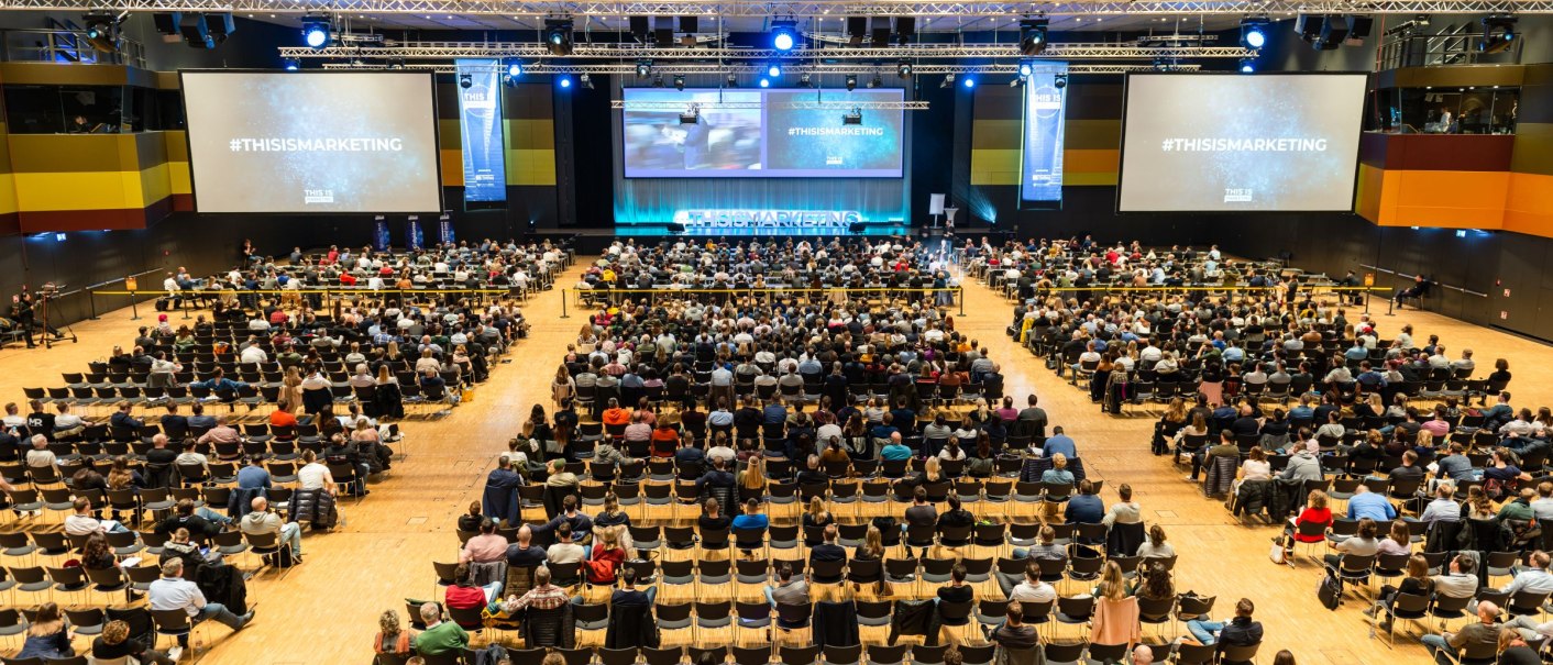 Gro&szlig;er Kongresssaal mit vielen Teilnehmern, die einer Pr&auml;sentation folgen. Zwei gro&szlig;e Bildschirme zeigen '#THISISMARKETING'. Die B&uuml;hne ist beleuchtet., &copy; Messe Stuttgart