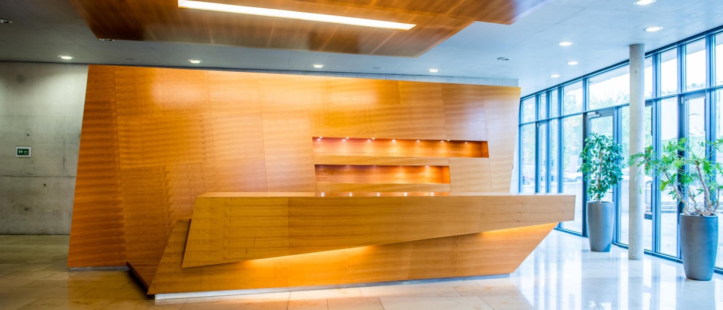 Modernes Foyer mit einer stilvollen Holztheke, beleuchtetem Regal und großen Fenstern. Helle Fliesen und Pflanzen in Töpfen schmücken den Raum., © GIACINTO_CARLUCCI