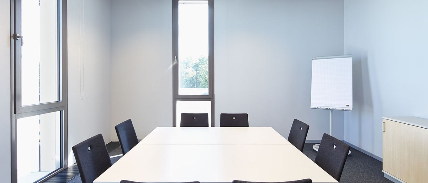 Ein moderner Seminarraum mit einem weißen Tisch, umgeben von schwarzen Stühlen. Ein Flipchart steht an der Wand, Fenster lassen Tageslicht herein., © Steffen Scheyhing Fotodesign