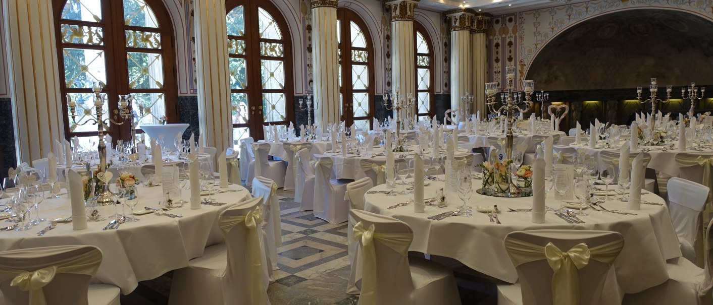 Eleganter Bankettsaal mit runden Tischen, weißen Tischdecken und Stuhlhussen. Große Fenster und dekorative Säulen verleihen dem Raum eine luxuriöse Atmosphäre., © marmorsaalgmbh