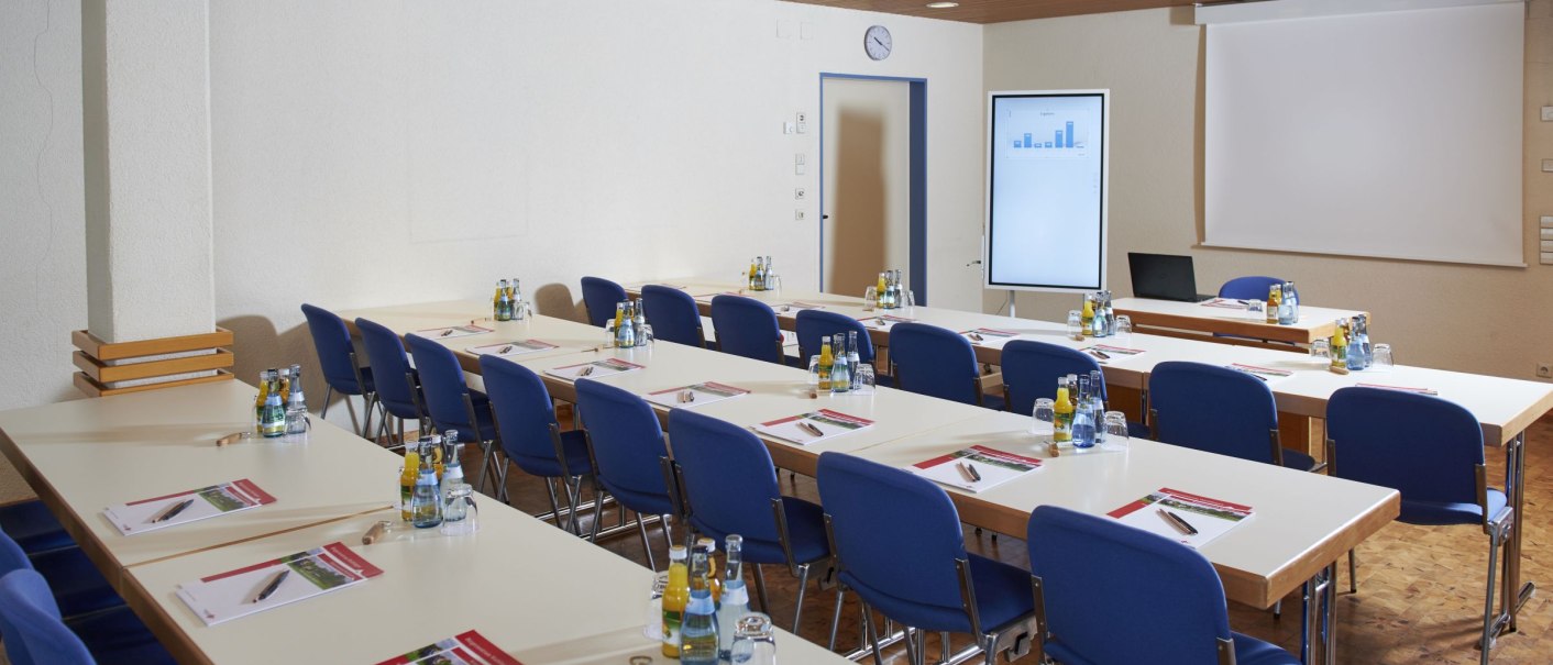 Konferenzraum mit blauen Stühlen und Tischen, ausgestattet mit Getränken und Schreibmaterial. Ein Bildschirm und eine Leinwand sind vorhanden., © Peter Oppenländer