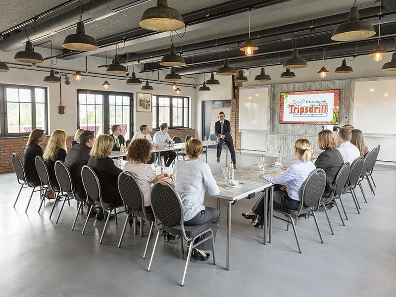 Ein moderner Konferenzraum mit Menschen, die an Tischen sitzen. Ein Bildschirm zeigt das Logo von Erlebnispark Tripsdrill., © Claudia Fy