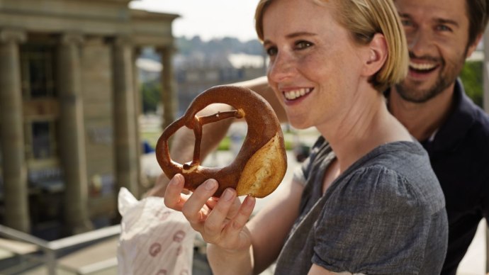 Frau hält eine schwäbische Brezel, lächelnder Mann im Hintergrund, Gebäude im Hintergrund., © TMBW, Christoph Düpper