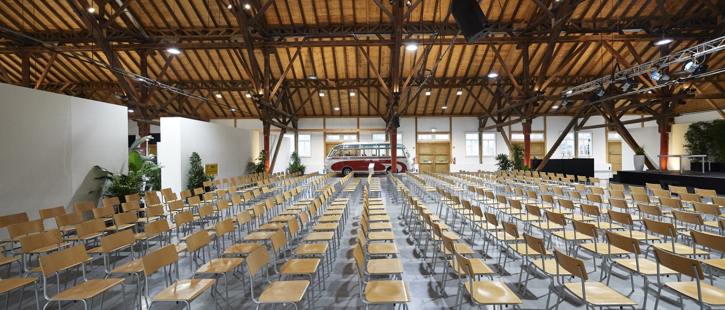 Gro&szlig;e Halle mit Holzdecke, leeren Stuhlreihen und einem roten Bus im Hintergrund. Pflanzen und Beleuchtung sind sichtbar., &copy; Peter Hartung